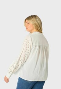 Blusa bianca con maniche lunghe ricamate, colletto arricciato e vestibilità ampia, abbinata a jeans blu. Tessuto morbido e leggero.