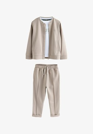 Beige jakke med lynlås og lange ærmer, matchende bukser og en hvid T-shirt nedenunder. Hvide sneakers med velcrolukning og lysebrune såler.