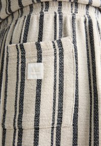 Tissu beige texturé avec des rayures noires verticales et une poche plaquée rectangulaire ornée d'une petite étiquette blanche.