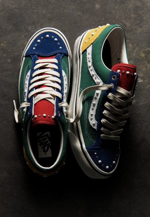 Deux baskets Vans colorées, à lacets blancs et à panneaux cloutés, sont posées sur une surface sombre.