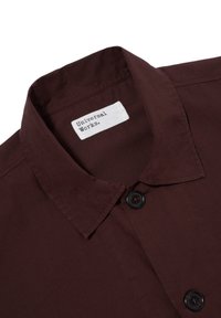 Camicia bordeaux con colletto a punta, bottoni neri e un'etichetta che riporta 'Universal Works'. Tessuto dalla consistenza morbida.
