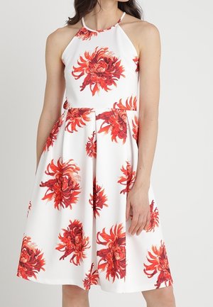 White sleeveless dress with large red floral patterns and a halter neckline worn by a standing woman against a plain background.