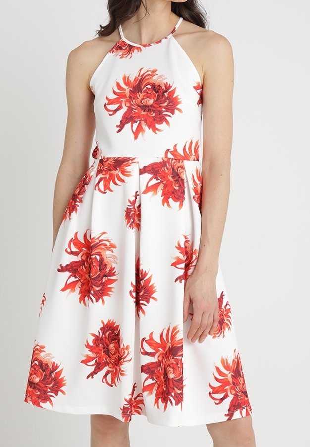 Robe blanche sans manches avec de grands motifs floraux rouges et un décolleté dos nu portée par une femme debout sur un fond uni.