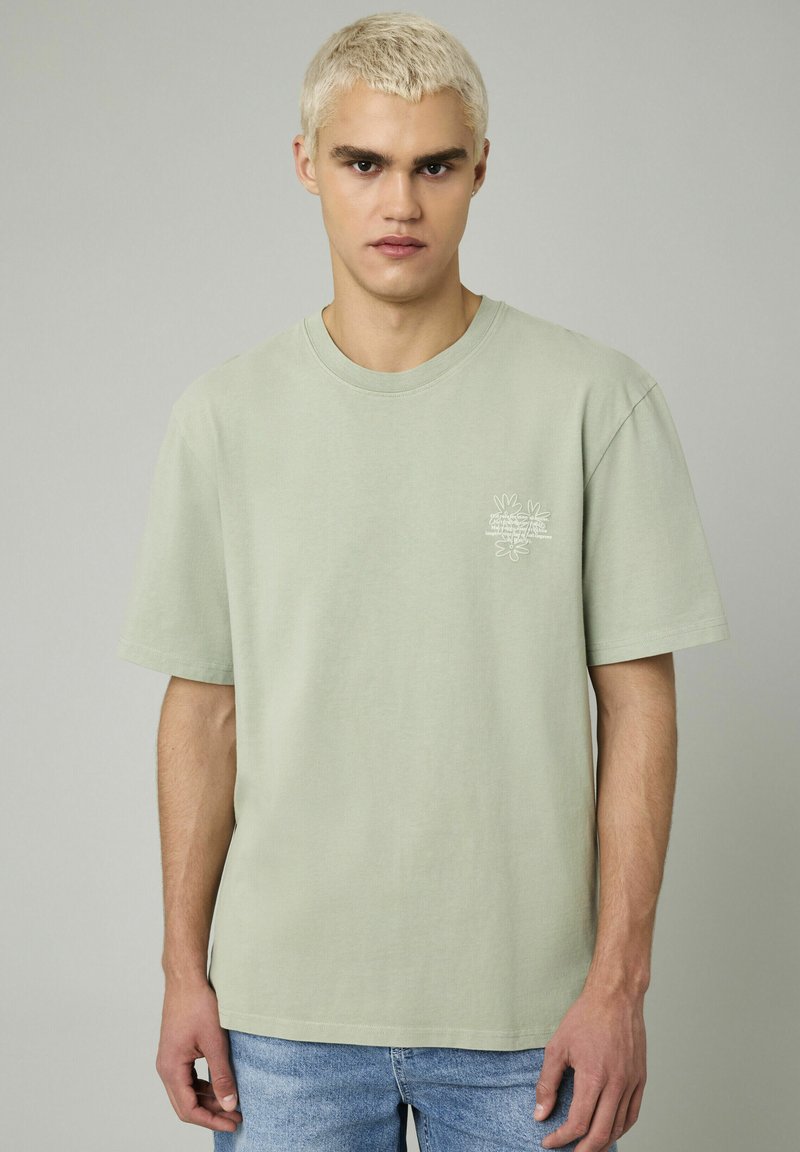 T-shirt en coton vert menthe avec un col rond et des manches courtes. Présente un petit design graphique blanc sur la zone gauche de la poitrine.