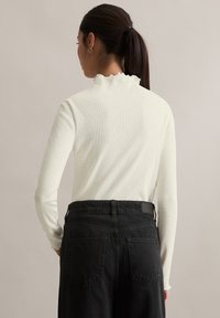 Kremowy top z długim rękawem i falbanianym kołnierzem, zestawiony z czarnymi dżinsami z wysokim stanem, które mają kieszenie z tyłu i gładki pas.