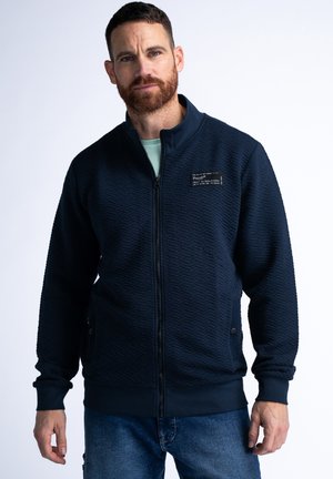 Marine blauwe ritsjack met textuurstof, ribgebreide mouwen en zoom, voorzien van een label op de borst en zijzakken. Katoen/polyester mix.