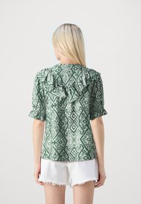 Blusa verde a fantasia con maniche volantee, caratterizzata da un design geometrico. Abbinata a pantaloni corti bianchi sfilacciati, vista posteriore.