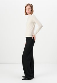 Woman wearing a fitted beige long-sleeve top and wide-leg black pants standing sideways with one hand on her hip against a white background.