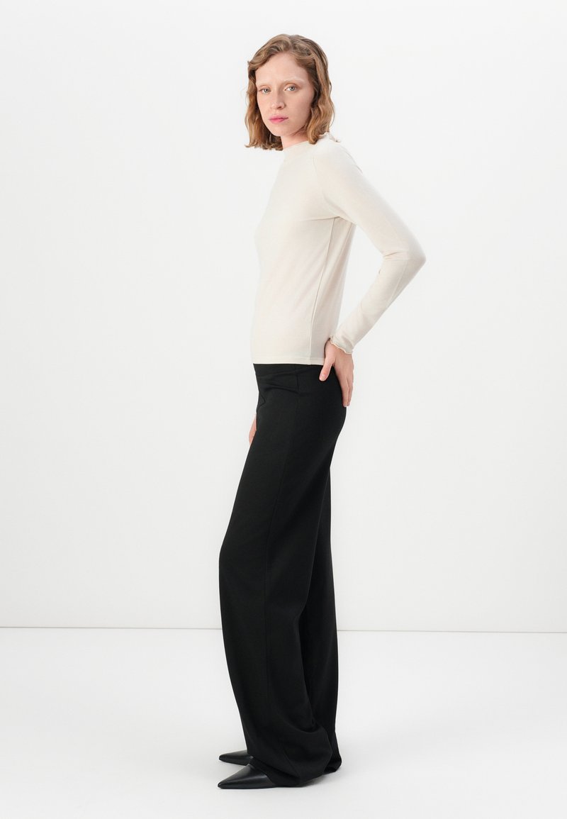 Donna che indossa un top lungo aderente beige e pantaloni neri a gamba larga, in piedi di lato con una mano sul fianco, su uno sfondo bianco.