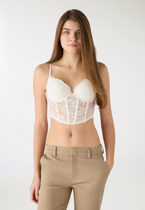 Giovane donna con capelli lunghi che indossa un bustier di pizzo bianco e pantaloni beige, in piedi con una mano in tasca su uno sfondo semplice.