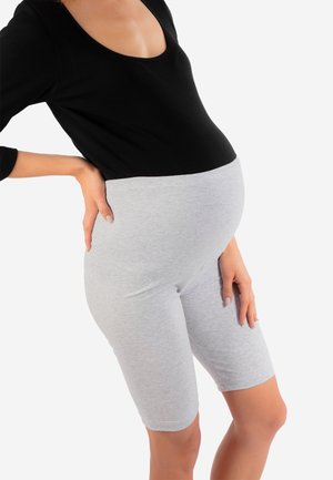 Osoba w ciąży ubrana w czarną bluzkę i jasnoszare szorty ciążowe, opierająca jedną rękę na dolnej części pleców, stojąca na białym tle.