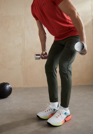 Mann in rotem Hemd und grüner Hose hebt zwei silberne Kurzhanteln in einem minimalistischen Innen-Fitnessstudio mit einem schwarzen Medizinball auf dem Boden.