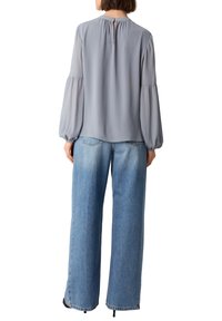 Grau, langärmlige Bluse mit gerafftem Kragen und elastischen Bündchen, kombiniert mit high-waisted hellblauen weit geschnittenen Jeans. Der Stoff wirkt leicht.