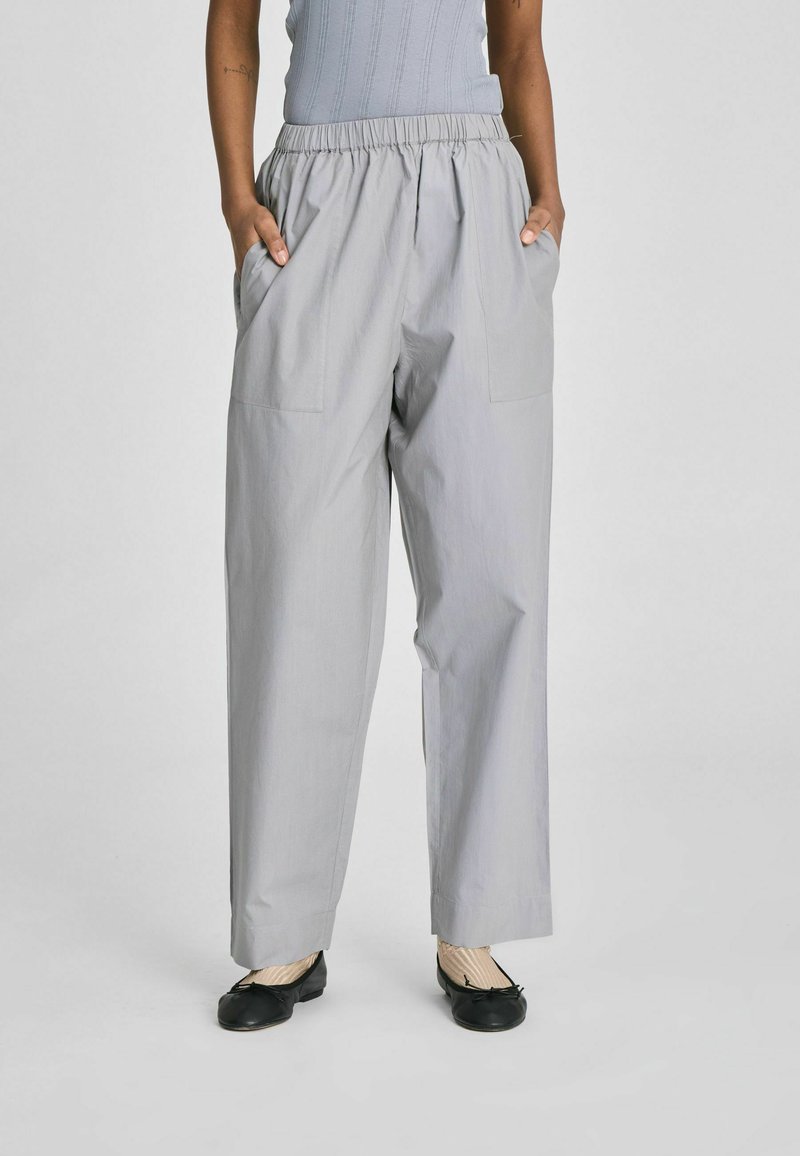 Személy világosszürke, laza szabású nadrágot visel gumis derékrésszel és nagy első zsebekkel, fekete balerinacipővel és krémszínű zoknival párosítva.