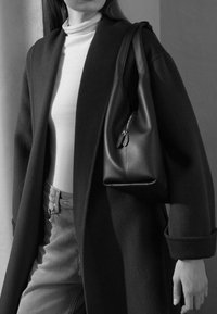 Borsa in pelle nera con dettaglio di cerniera, indossata sopra un dolcevita bianco e un cappotto scuro, abbinata a jeans grigio chiaro. Caratteristiche di design minimalista.