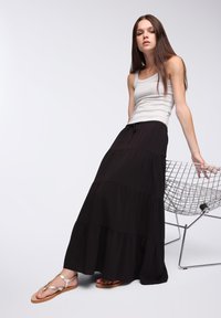 Czarna, warstwowa spódnica maxi z regulowanym pasem, w połączeniu z szarym topem na ramiączkach i metalicznymi sandałami, ustawiona obok metalowego krzesła.