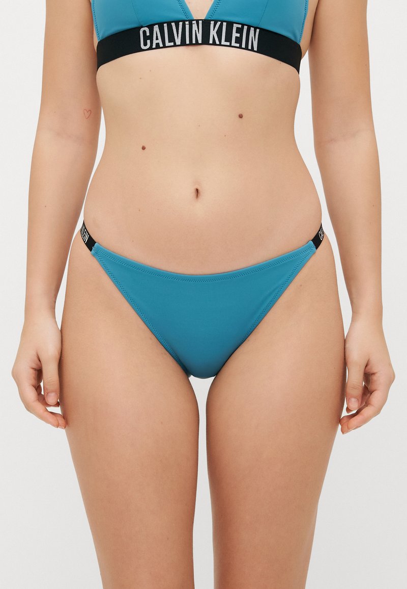 Tyrkysový bikiny set s braletou ve tvaru V a nízkými kalhotkami. Obsahuje logo pás s černobílými akcenty, vyrobeno z hladkého materiálu.