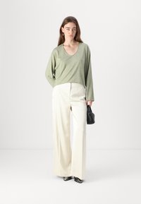 Ljusgrön långärmad topp med V-ringning som matchas med vita vida byxor. Modellen håller en svart handväska. Slät tygtextur.