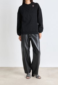 Sweatshirt nero con logo a rilievo, abbinato a pantaloni larghi neri lucidi e sandali con tacco a blocco decorati da un motivo e testo.