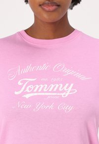 Růžové bavlněné tričko s bílým tištěným textem: "Autentické originální Tommy jeans, založeno 1985, New York City." Kulatý výstřih, krátké rukávy.
