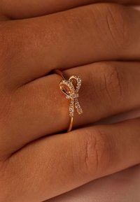 Bague en or au design en nœud, ornée de petites pierres scintillantes, posée sur un anneau lisse, présentée sur une main devant un fond clair.