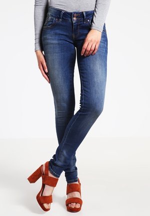 Jeans ajustados en denim azul oscuro con detalles deslavados, presentan un cierre de botón y bolsillos frontales. Combinados con sandalias de plataforma de ante marrón.