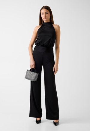 Donna che indossa un top nero senza maniche con collo alto e pantaloni a gamba larga, che tiene in mano una piccola borsa a quadri bianca e nera su uno sfondo semplice.