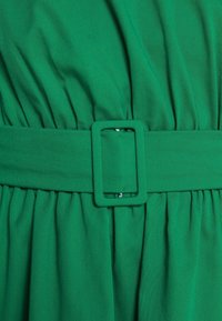 Grön klänning med ett brett midjeband och en rektangulär bältesspänne, plisserad textur längs midjan och ett mjukt, flödande tyg.