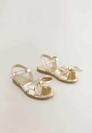 Auksinės metalinės basutės su reguliuojamu kulkšnies dirželiu, priekyje įrišta kaspinėlio detalė ir plokščia padais. Išgaubtas paviršius visame paviršiuje.