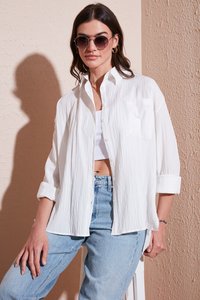 White, textured button-up shirt with a single chest pocket, paired with light blue jeans. Model wearing round sunglasses.