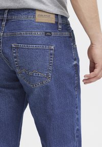 Jeans de mezclilla azul con un acabado texturizado, que presentan un parche de cuero marrón, costuras en contraste y un diseño de bolsillo distintivo con un patrón en zigzag.