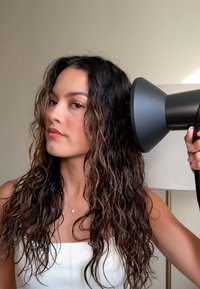Secador de pelo negro con una boquilla ancha, sostenido de lado cerca de cabello mojado y ondulado. Modelo con un top blanco sin tirantes, fondo claro.