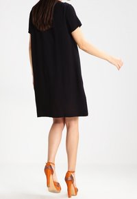 Robe noire à manches courtes, coupe décontractée, arrivant à mi-cuisse. Portée avec des chaussures à talons hauts orange dotées de brides transparentes et d'accents décoratifs.