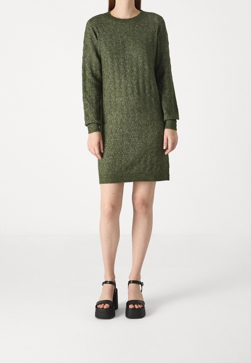 Femme portant une robe pull tricotée à torsades longueur genoux vert olive et des sandales à plateforme noires avec des brides à la cheville sur un fond uni.