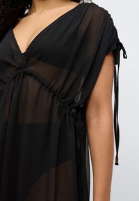 Μαύρο, διάφανο φόρεμα με ντεκολτέ σε σχήμα V, σικερισμένη μέση και ρυθμιζόμενα κορδόνια στους ώμους. Ελαφρύ, υφασμάτινο με υφή και απαλό πέπλο.