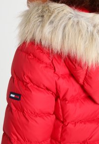 Czerwona kurtka ocieplana z kapturem wykończonym futrem. Posiada pikowaną teksturę i naszywkę z logo na rękawie. Materiał wydaje się gładki i błyszczący.