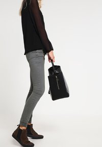 Czarna torba o żebrowanej strukturze, trzymana przez osobę w czarnej bluzce i szarych obcisłych dżinsach, noszącą brązowe botki za kostkę z czarnymi akcentami.