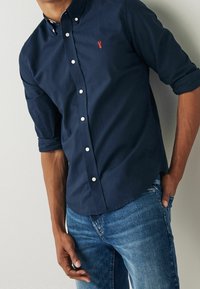 Chemise boutonnée en coton bleu marine avec un petit logo de homard rouge, dotée d'un col et de manches retroussées, associée à un jean en denim bleu.