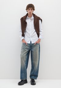 Gilet lavorato a maglia marrone sopra una camicia bianca a righe con bottoni, abbinato a jeans larghi azzurri sdruciti e scarpe platform nere.