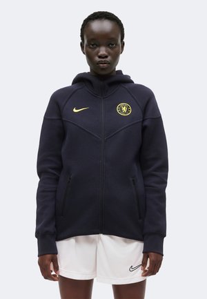Isik, kes kannab musta Chelsea jalgpalli lukuga huppi kollaste logodega ja valgeid Nike lühikesi pükse, seisab ühevärvilise tausta ees.