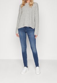 Suéter gris de punto con un corte relajado, combinado con jeans ajustados azules y zapatillas blancas. Los detalles notables incluyen un dobladillo deshilachado en los jeans.