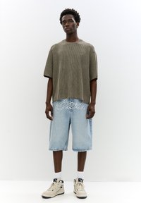 Oliven grøn, ribbet strik t-shirt med korte ærmer, parret med lyseblå denim shorts med hvidt broderet tekst, og beige sneakers.
