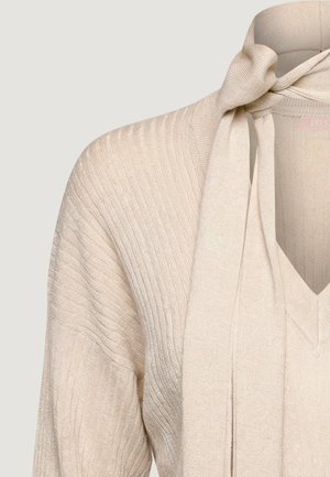 Pull en maille côtelée beige avec col en V et écharpe de cou attachée nouée sur un fond uni clair.