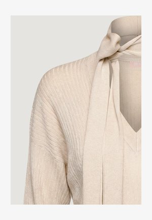 Beżowy prążkowany sweter z dekoltem w serek i przymocowanym szalikiem przy szyi zawiązanym na supeł na jednolitym jasnym tle.