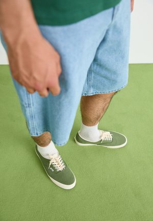 Žalios drobės sportbačiai su kreminiais raišteliais, dėvimi su baltais kojinėmis. Šviesiai mėlyni džinsiniai šortai, siekiantys šiek tiek virš kelių. Kambario grindys su tekstūra žalia spalva.