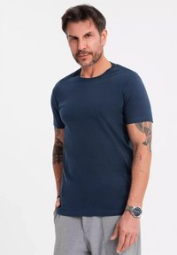 T-shirt blu navy a maniche corte realizzato in tessuto morbido, con un classico collo a giro. Tatuaggi visibili sul braccio e un orologio al polso.