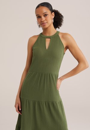 Naine kannab varrukateta oliivrohelist tekstuuri kleiti võtmeauguga kaelusega, poseerib tavalise beeži tausta ees.