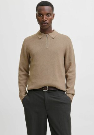 Hombre vestido con un suéter de cuello con cremallera de manga larga beige y pantalones gris oscuro, con las manos en los bolsillos, de pie frente a un fondo claro y liso.
