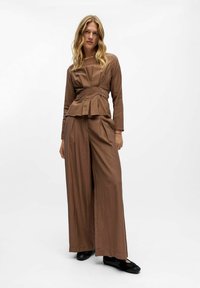 Femme aux longs cheveux blonds portant un chemisier plissé marron et un pantalon large, se tenant avec une main dans la poche et des chaussures noires.