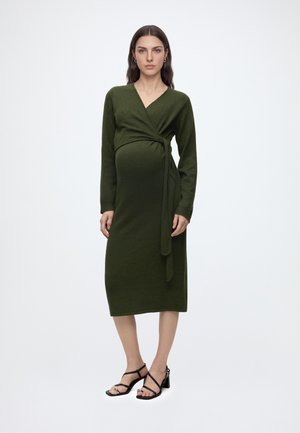 Femme enceinte portant une robe portefeuille à manches longues vert foncé et des sandales à talons noires, debout devant un fond clair uni.